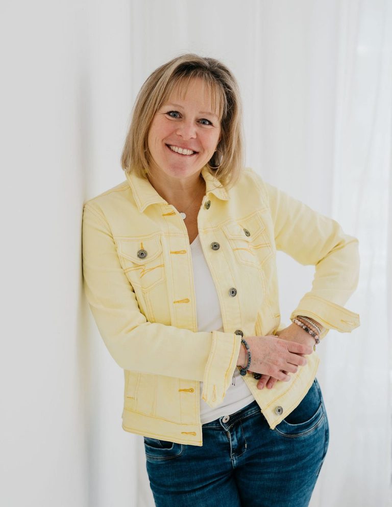 Eva Niggl Kinesiologie & Öle - Begleitung zu innerer Kraft und Stärke Lächelnde Frau mit leicht gewelltem Haar, stützt sich auf einen Tisch. Sie sieht freundlich und zufrieden aus.